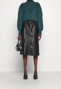 Pull en maille turquoise avec partie supérieure texturée, jupe midi en cuir noir avec poches sur les côtés, et chaussures à talons hauts noires. Sac noir à main.