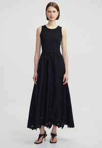 Schwarzes Maxikleid mit ärmellosem Design, geraffter Taille und Saum mit aufwendigen Lochmustern. Getragen mit hochhackigen Sandalen.