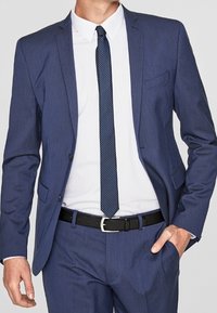 Mörkblå kostym med en slim fit-design, en knapp jacket, snedskurna slag och ett strukturerat slips. Svart bälte med en silvrig spänne.