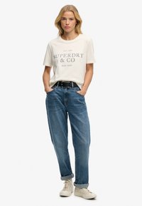 Roomkleurig T-shirt met korte mouwen en grijze tekst, gecombineerd met relaxte blauwe spijkerbroek. Witte sneakers, zwarte riem. Simpele, casual outfit.