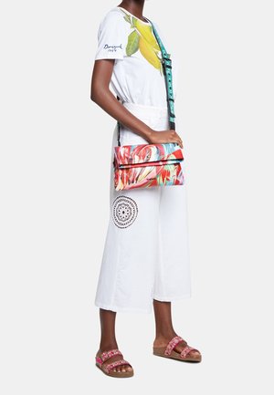 Barevná crossbody taška s vířivým květinovým vzorem, černými akcenty a nastavitelným popruhem. Spárovaná s bílými širokými kalhotami.