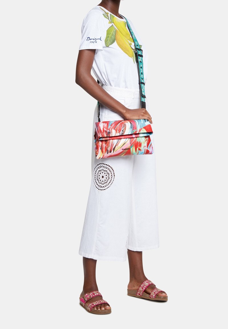 Kolorowa torba na ramię z wirującym kwiatowym wzorem, czarnymi akcentami i regulowanym paskiem. W połączeniu z białymi spodniami z szerokimi nogawkami.