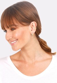Pendientes en tono dorado con una combinaci�ón de aros y tachuelas. Presentan gemas transparentes y un acabado texturizado, exhibidos sobre colores neutros.