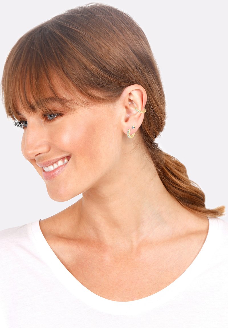 Pendientes en tono dorado con una combinación de aros y tachuelas. Presentan gemas transparentes y un acabado texturizado, exhibidos sobre colores neutros.