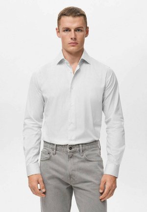 Camisa blanca de manga larga con botones y un sutil patrón a rayas, combinada con jeans gris claro. Presenta un cuello clásico y puños abotonados.
