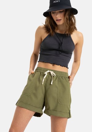 Olivgrüne Shorts mit umgeschlagenen Säumen und Seitentaschen, kombiniert mit einem schwarzen gerippten Crop-Top und einem schwarzen Bucket Hat mit dem Markennamen.