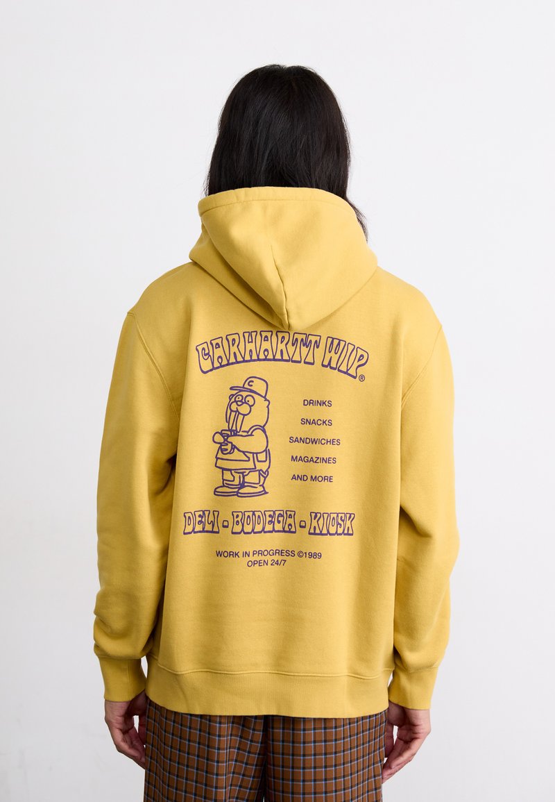 Gelbe Kapuzenjacke mit lila grafikdruck. Zeigt eine Cartoonfigur, die Lebensmittel hält, der Text enthält "Deli Bodega Kiosk" und die dazugehörigen Dienstleistungen.