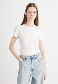 Camiseta de algodón blanca con mangas cortas, cuello redondo y un pequeño logo en el pecho, combinada con jeans de tiro alto de color azul claro.