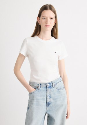 T-shirt bianca in cotone con maniche corte, colletto a giro, e un piccolo logo sul petto, abbinata a jeans a vita alta di colore blu chiaro.