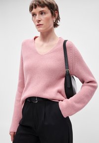 Ροζ ριμπ πλεκτό πουλόβερ με V λαιμόκοψη, διαθέτει μακριά μανίκια και χαλαρή εφαρμογή, σε συνδυασμό με εφαρμοστά μαύρα παντελόνια και μια μαύρη τσάντα ώμου.