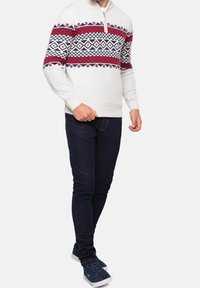 Suéter gris claro con media cremallera, patrones geométricos rojos y marinos en el pecho, combinado con vaqueros azul oscuro y zapatillas marinas.