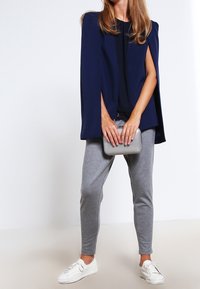 Blazer estilo capa en azul marino sobre una blusa negra, combinado con pantalones ajustados grises y zapatillas blancas. Sosteniendo un clutch plateado. Tejido suave y corte a medida.