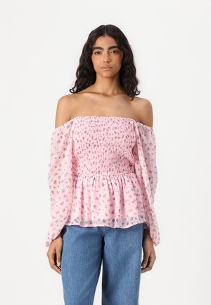 Schulterfreie Bluse im pinken Blumenmuster, mit gerafftem Oberteil und transparenten, puffigen langen Ärmeln. Kombiniert mit blauen Jeans.