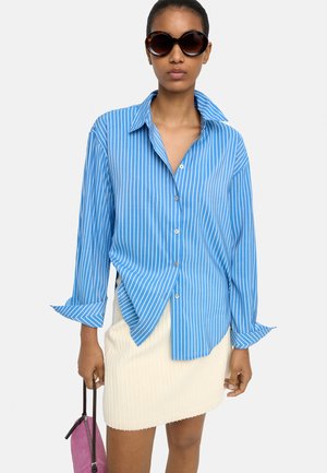 Kobieta w oversize niebiesko-białej koszuli w paski, kremowej plisowanej spódnicy, dużych okrągłych okularach przeciwsłonecznych, trzymająca małą różową torebkę.