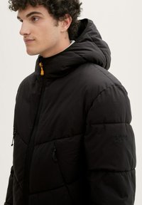 Veste doudoune noire avec une fermeture éclair à l'avant, deux poches latérales, une capuche et un léger éclat. Comprend des cordons de serrage ajustables et un logo texturé sur la manche.