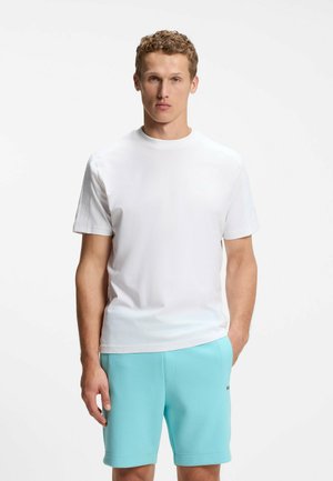Giovane uomo in piedi di fronte, indossa una t-shirt bianca semplice e pantaloncini azzurri con una mano in tasca.
