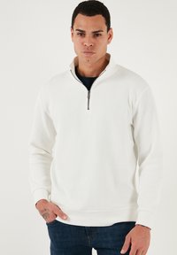 Biely rebrovaný pulóver so štvrťzipom a dlhými rukávmi. Obsahuje pevnú pletenú textúru a priliehavé manžety.