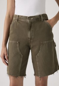 Γυναίκα που φοράει χακί πράσινα τζιν σορτς μήκους ως το γόνατο με αδέξια τελειώματα και μεγάλες μπροστινές τσέπες, συνδυασμέν α με λευκό ριμπ τοπ χωρίς μανίκια.