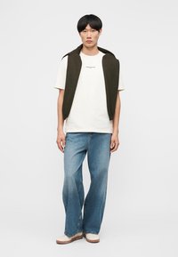 Νεαρός άνδρας που φοράει λευκό T-shirt, μπλε φαρδιά τζιν παντελόνια, λευκά παπούτσια και ελιά πουλόβερ ριγμένο στους ώμους του.