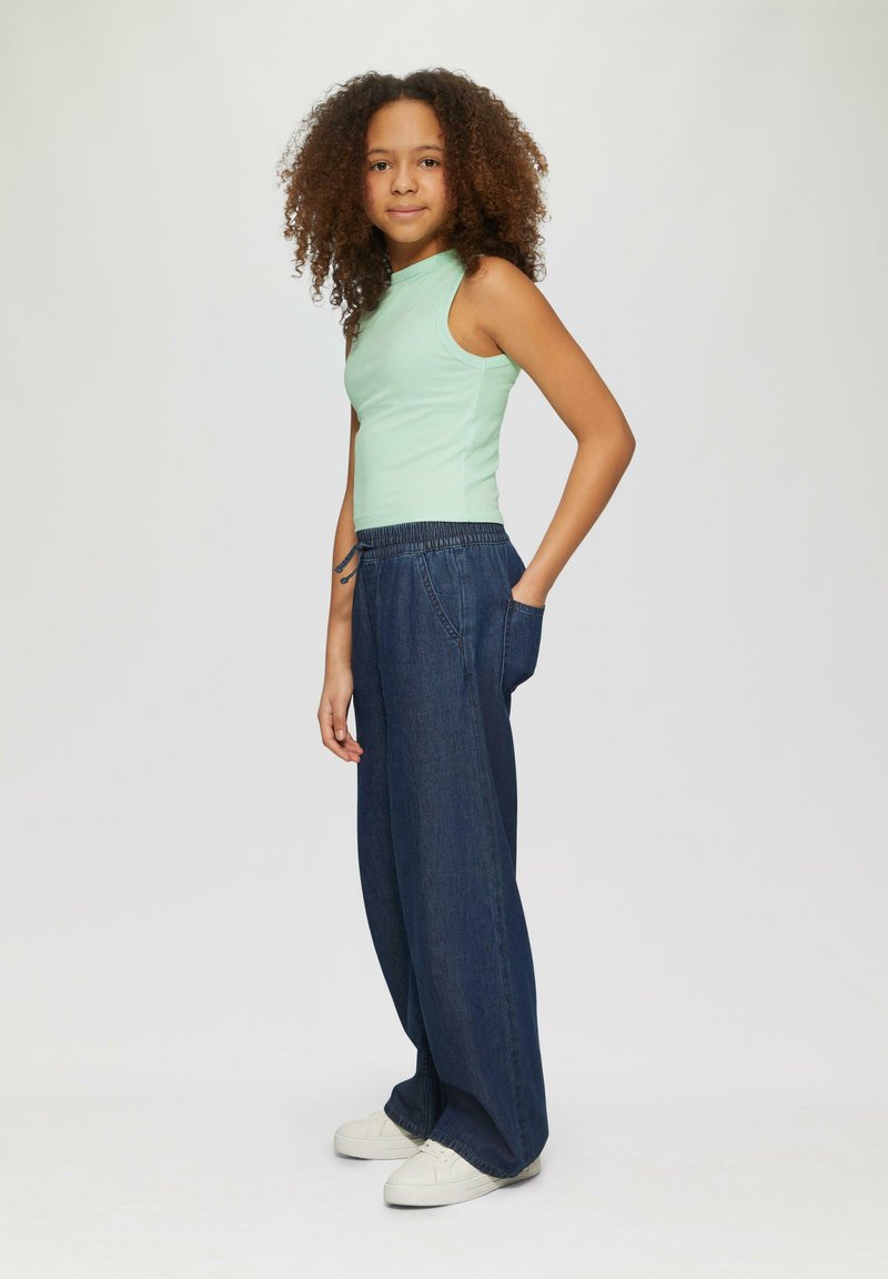 Hellgrünes, geripptes, ärmelloses Oberteil, kombiniert mit dunkelblauen, weit geschnittenen Jeans mit elastischem Bund und Taschen, getragen mit weißen Sneakers.