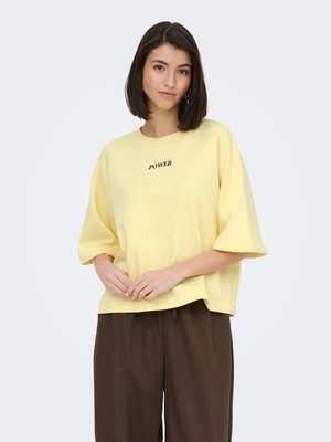 Femme aux cheveux lisses et foncés portant une chemise ample jaune pâle avec le texte « POWER » et un pantalon large marron, debout devant un fond clair.