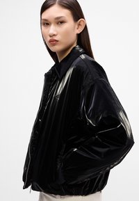 Jachetă bomber din nailon negru cu un finisaj lucios, închidere cu fermoar și guler. Dispune de manșete elastice și o croială confortabilă.