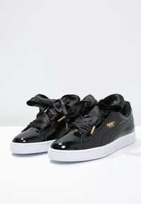 Baskets en cuir verni noir avec semelle en caoutchouc blanc, lacets en ruban de satin noir et accents de logo doré sur le côté.