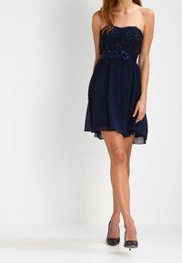 Mörkblå axelbandslös klänning med spetsliv, flödande kjol och rosett vid midjan. Slät yta, knälång med genomskinlig överlag.