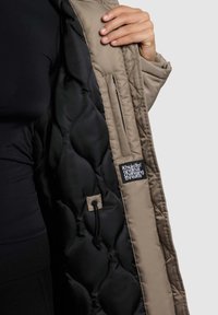 Beež värvusega polsterdatud jope, millel on must tikandiga vooder logoga sildiga. Siseküljel on reguleerimiseks tõmbeköis.