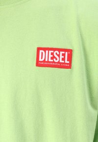 Zelené bavlněné tričko s červeným obdélníkovým logem, na kterém je bíle napsáno "DIESEL" a "PRO ÚSPĚŠNÝ ŽIVOT".