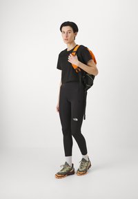 Black cropped t-shirt, high-waisted black leggings, olive green and orange trail shoes, and an orange backpack with black accents.