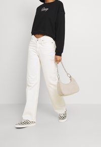Felpa nera corta con ricamo "Roxy", jeans bianchi a gamba larga, scarpe slip-on a scacchi bianco e nero, e borsa beige con tracolla a catena.