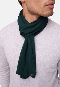 Mørkegrøn strikket tørklæde draperet om halsen, med ribbet struktur og et lille sort mærke i enden, sat sammen med en lys grå sweater.