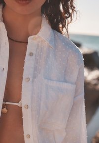 Camicia bianca con pois in rilievo, bottoni sul davanti e taschino sul petto, indossata sopra un top bikini bianco. Luce naturale e sfondo marino.