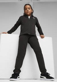 Melns zip-aizdarejams iedobums ar PUMA logo, apvienots ar atbilstošiem sporta biksēm, abiem izgatavotiem no mīksta materiāla. Melni tenisi ar baltām zolem.