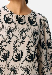 Blouse beige avec broderie florale noire présentant de grandes formes de pétales et des motifs de vignes tourbillonnants, portée par une personne avec le cou et les épaules visibles.