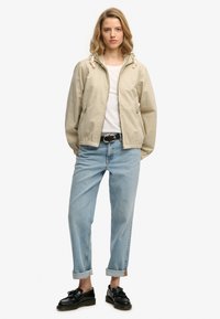 Chaqueta beige claro con capucha, puños elásticos, cierre de cremallera, dos bolsillos laterales, combinada con vaqueros azul claro remangados y mocasines negros.