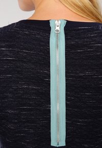 Musta paita, jossa on tekstuuri ja keskellä akvamarinvärinen vetoketju kaulasta helmaan, jota täydentää metallinen tarvikkeet.