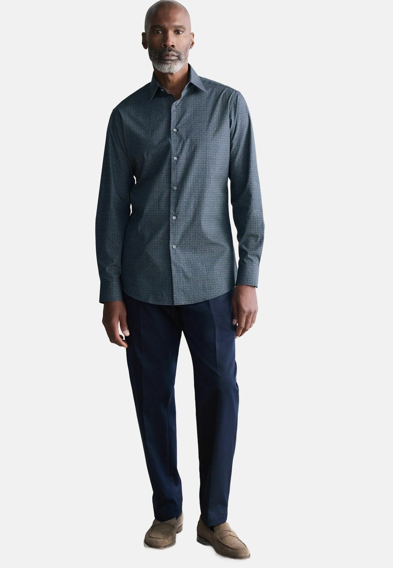 Camisa de botones de manga larga en azul oscuro con un patrón geométrico; combinada con pantalones navy y zapatos de ante marrón claro.