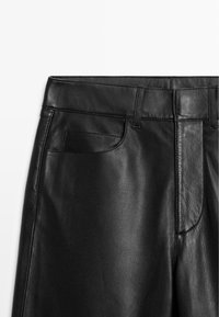 Schwarze Lederhosen mit glatter Textur, traditionellem Taillenbund und vorn liegenden Taschen. Nähte sind entlang der Säume sichtbar.