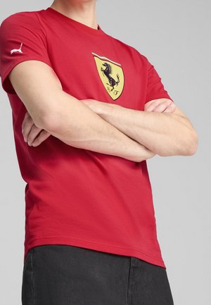 Rotes Baumwoll-T-Shirt mit einem gelben Schild-Logo, auf dem ein schwarzes Pferd abgebildet ist, kurzen Ärmeln und einem Rundhalsausschnitt. Modell trägt dunkle Hosen.