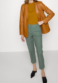 Bruine leren blazer, mosterdkleurig top en groen geruite broek, aangevuld met zwarte puntige muiltjes. Perfecte pasvorm, slank ontwerp.