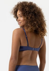 Námořnická modrá podprsenka s nastavitelnými ramínky a zapínáním na háček a oko. Hladký materiál; ramínka mají květinový vzor. Zvýrazněný pohled ze zad.