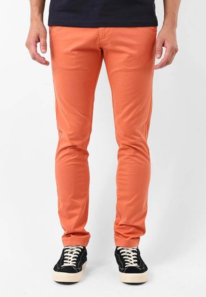 Pantalons slim orange en tissu extensible, avec une texture lisse et des détails de couture minimaux. Associés à des baskets noires.