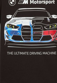 Juodas marškinėliai su BMW automobilio grafika, kuri turi mėlynus, baltus ir raudonus akcentus; po grafika yra užrašas "GALUTINIS VAŽIAVIMO ĮRANGOS".