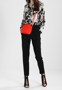 Kobieta w czarnej kwiecistej bluzce, czarnych spodniach z wysokim stanem, czarnych sandałach na obcasie z odkrytymi palcami, trzymająca jasnoczerwoną torebkę na ramię.