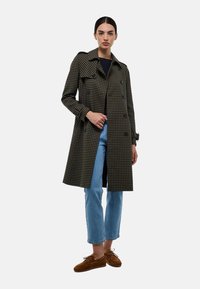 Manteau trench noir et vert à carreaux avec un design croisé, épaulettes et manches longues, associé à un jean bleu clair et des chaussures marron.