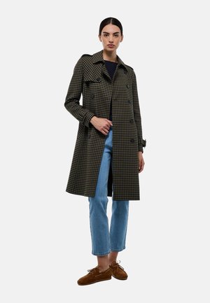 Manteau trench noir et vert à carreaux avec un design croisé, épaulettes et manches longues, associé à un jean bleu clair et des chaussures marron.
