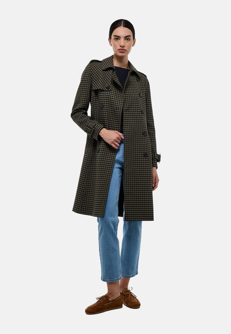Manteau trench noir et vert à carreaux avec un design croisé, épaulettes et manches longues, associé à un jean bleu clair et des chaussures marron.
