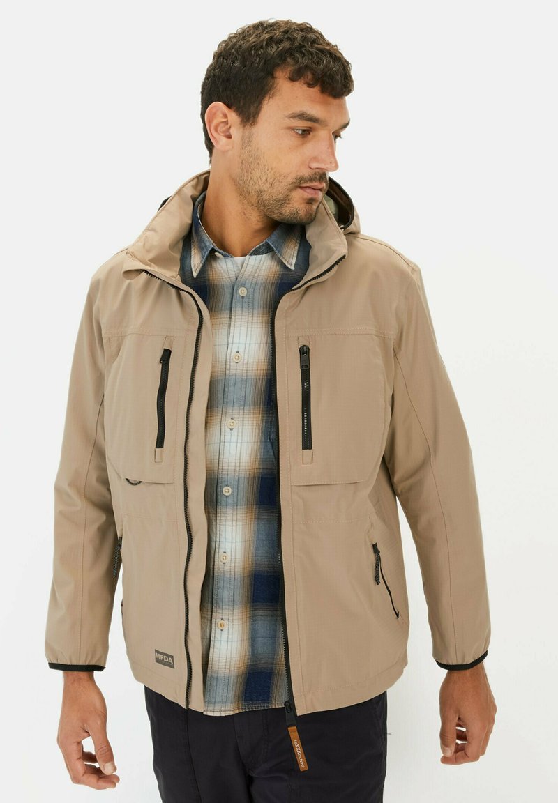 Beige waterdichte jas met een capuchon, ritszakken en verstelbare manchetten, gedragen over een blauw-bruin geruit shirt. Gladde textuur.